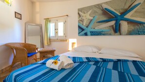 Habitació d'hotel amb llit, cadira, finestra amb cortines, làmpada penjada a la paret i quadre d'estries de mar.