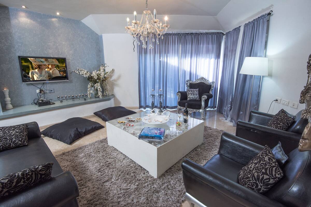 A modern living room with a glass coffee table, black leather sofas, a chandelier, and a television mounted on the wall.