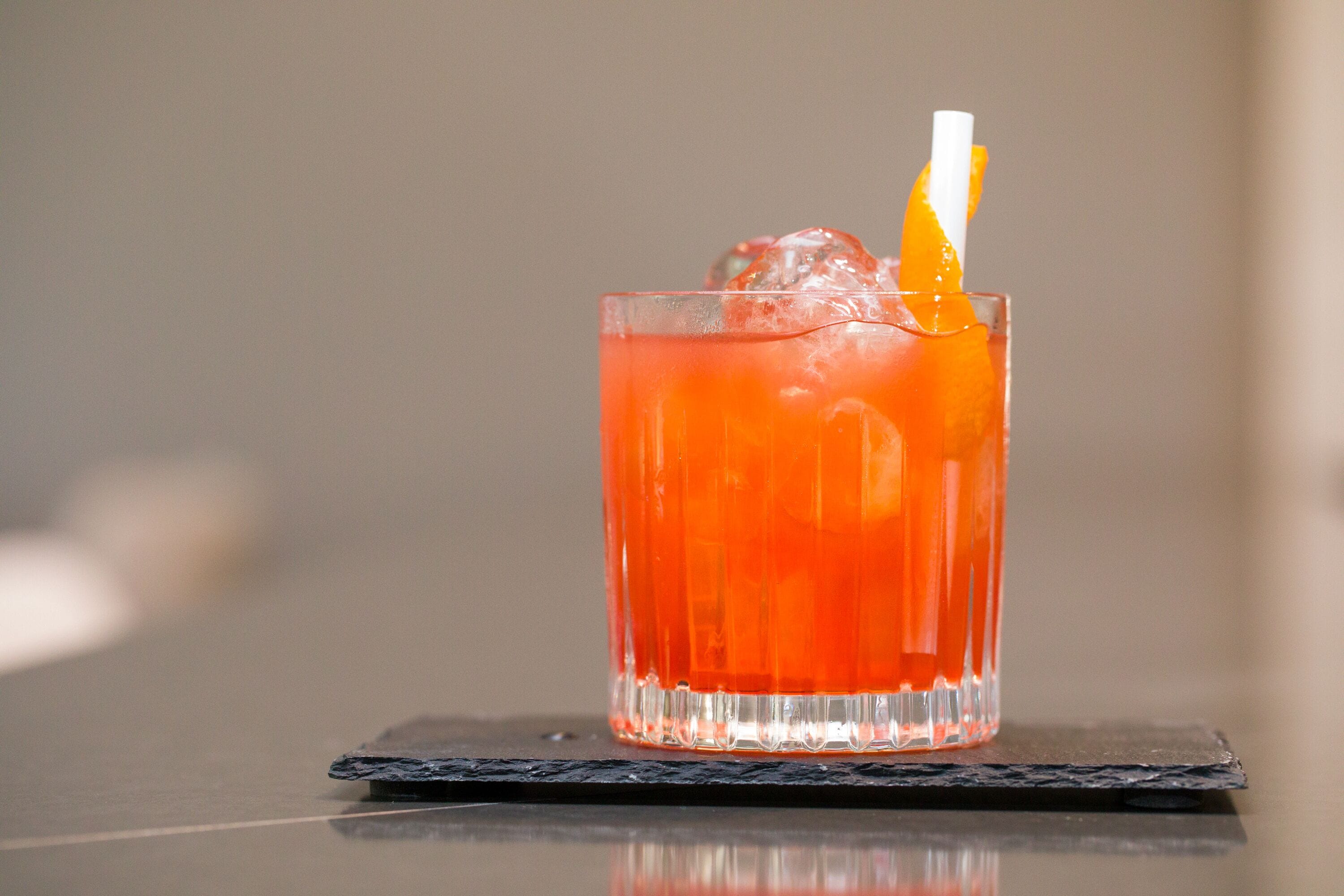A glass of red beverage with an orange slice and a straw on a black coaster.