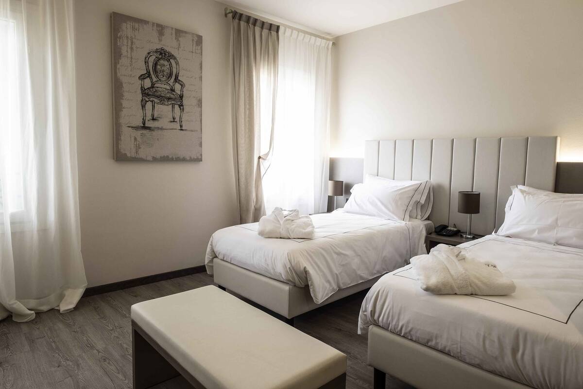 Hotellihuoneessa on kaksi sänkyä, penkki, yöpöytä lampun kanssa ja taulu seinällä.