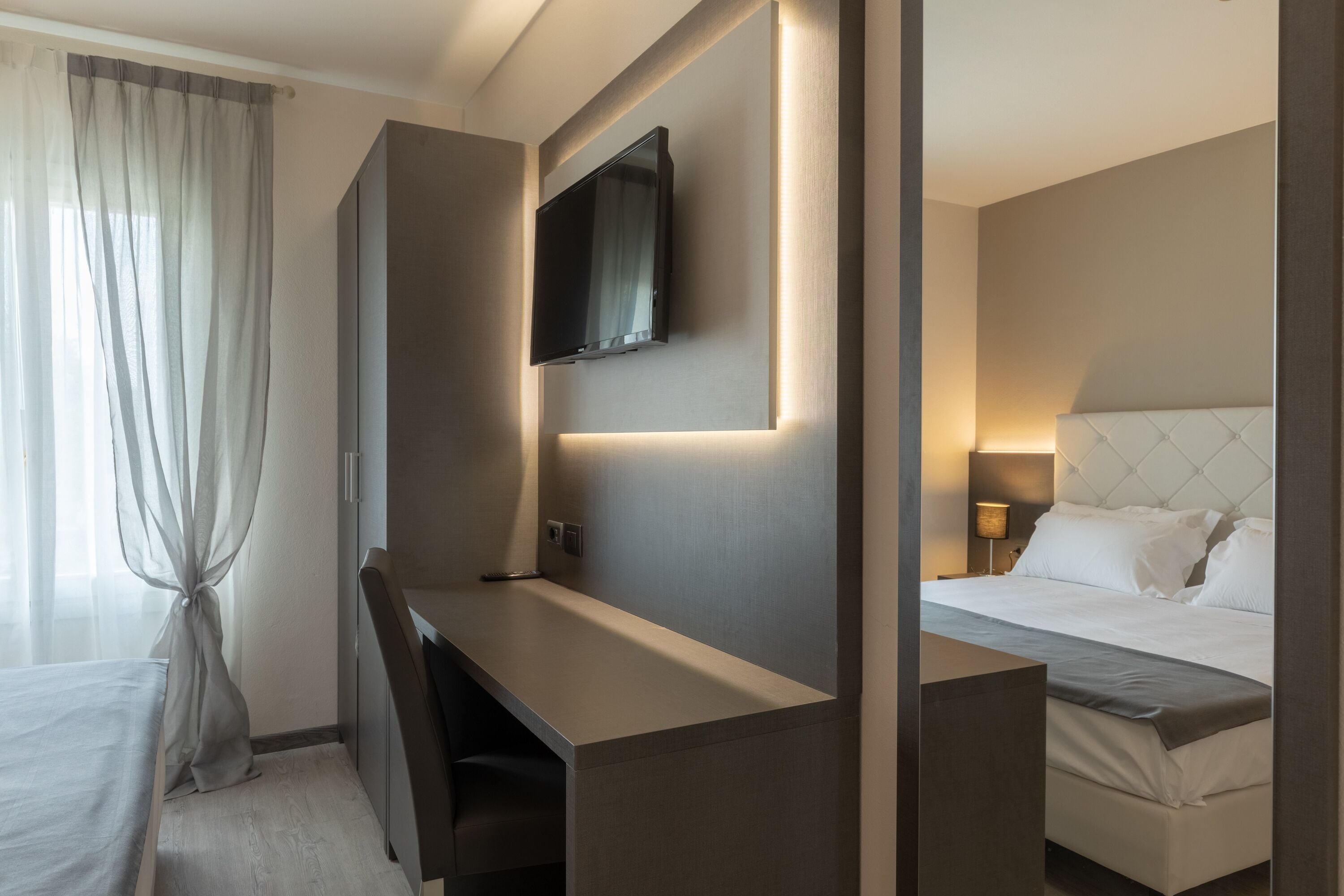 Una habitación de hotel moderna con cama, escritorio, televisor montado en la pared y ventana con cortinas de gasa.