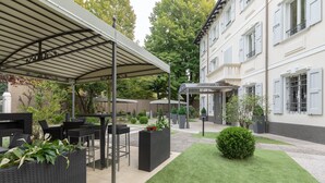 Een terras buiten met zwarte wicker meubels, een overdekte zithoek en een verzorgd gazon.
