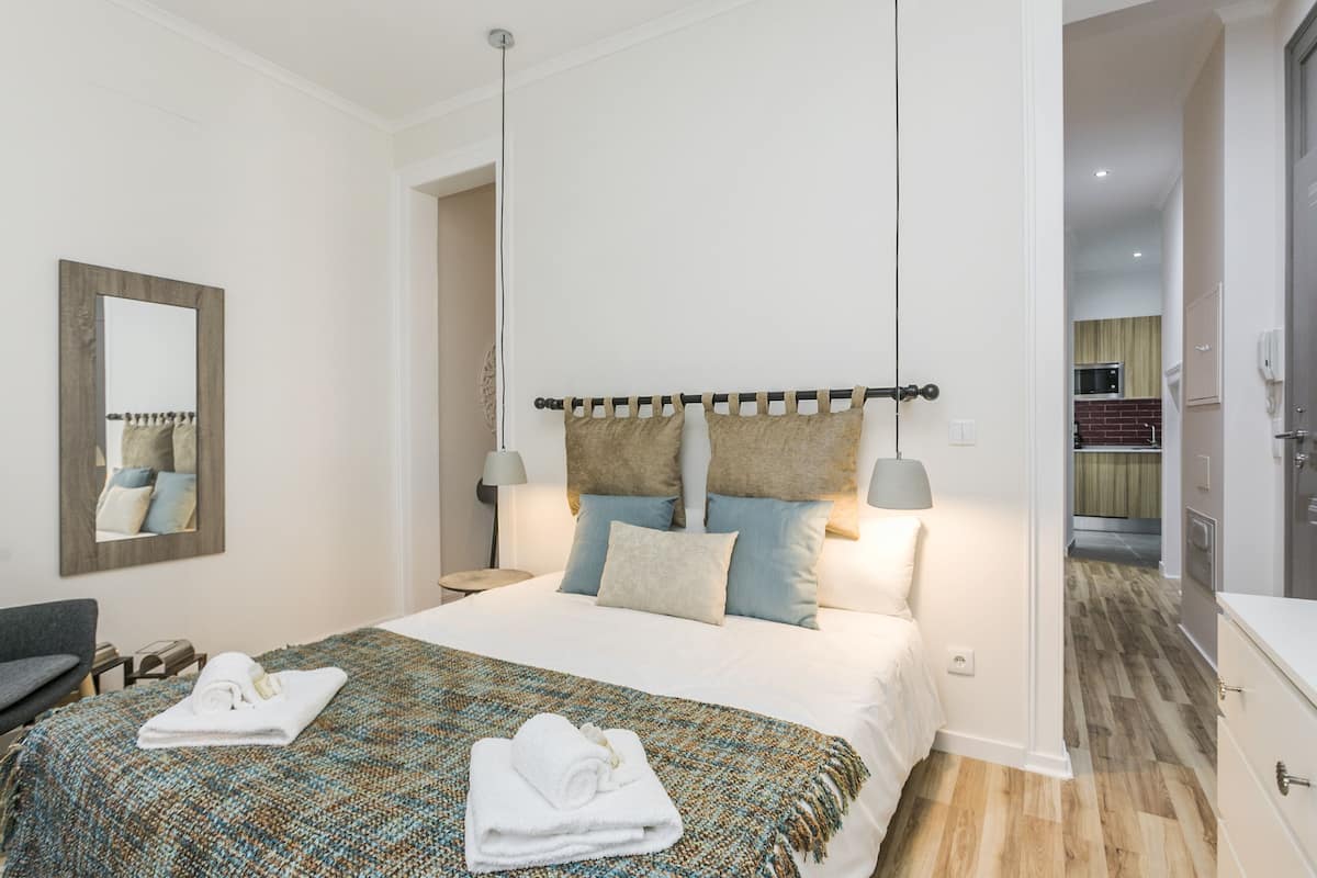 A modern bedroom with a large bed, a bedside table, a mirror, and a view into the kitchen.