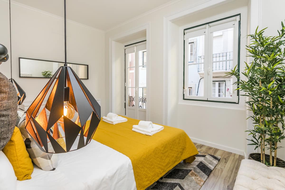 A hotel room with a bed, a hanging light fixture, a window with a view of buildings, and a potted plant.