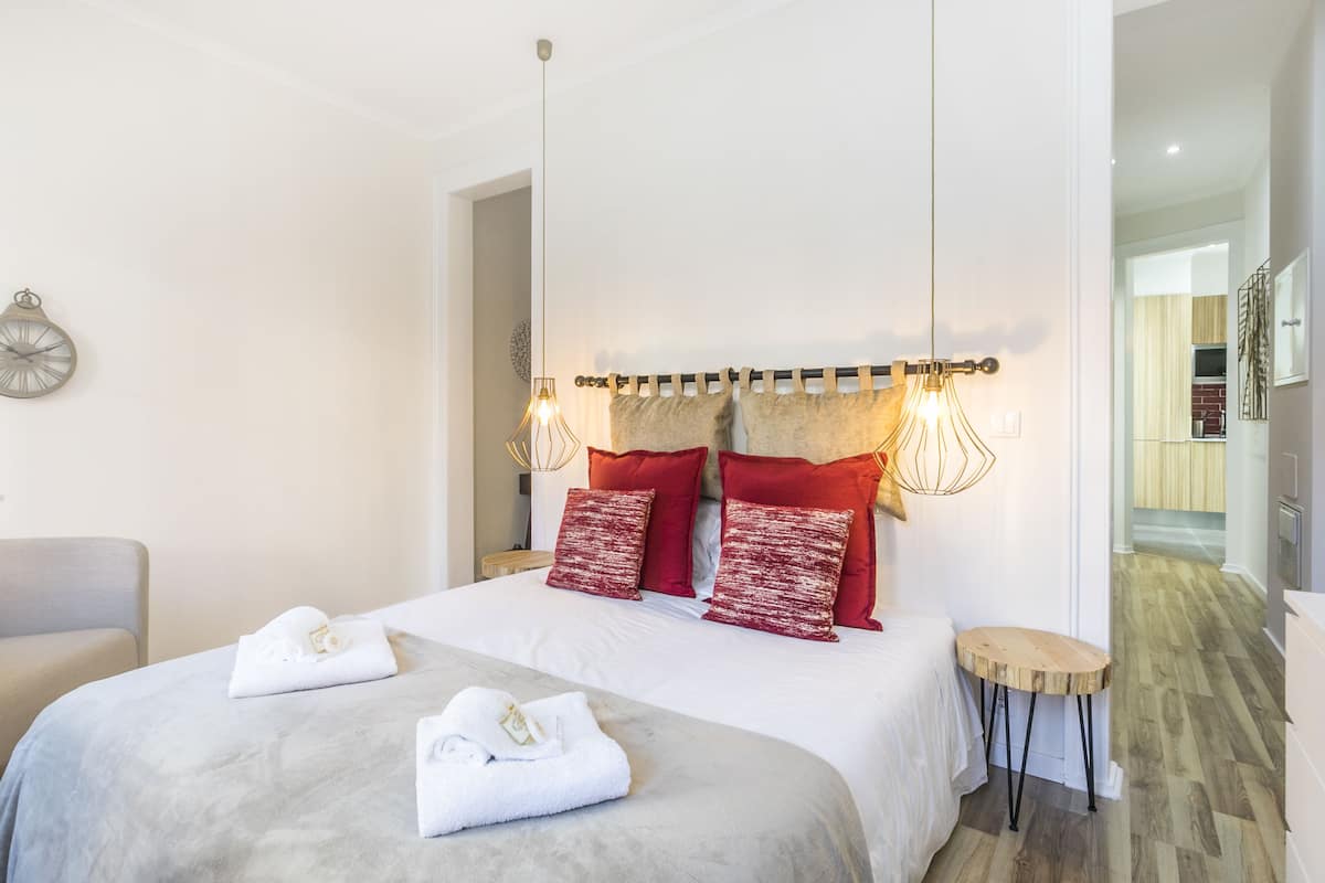 A hotel room with a large bed, two red pillows, a wooden side table, and a wall clock.