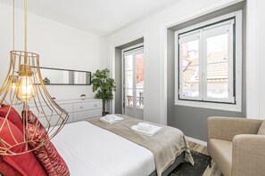 Une chambre d’hôtel avec un lit, une chaise, une plante, une commode et une fenêtre donnant sur des immeubles.