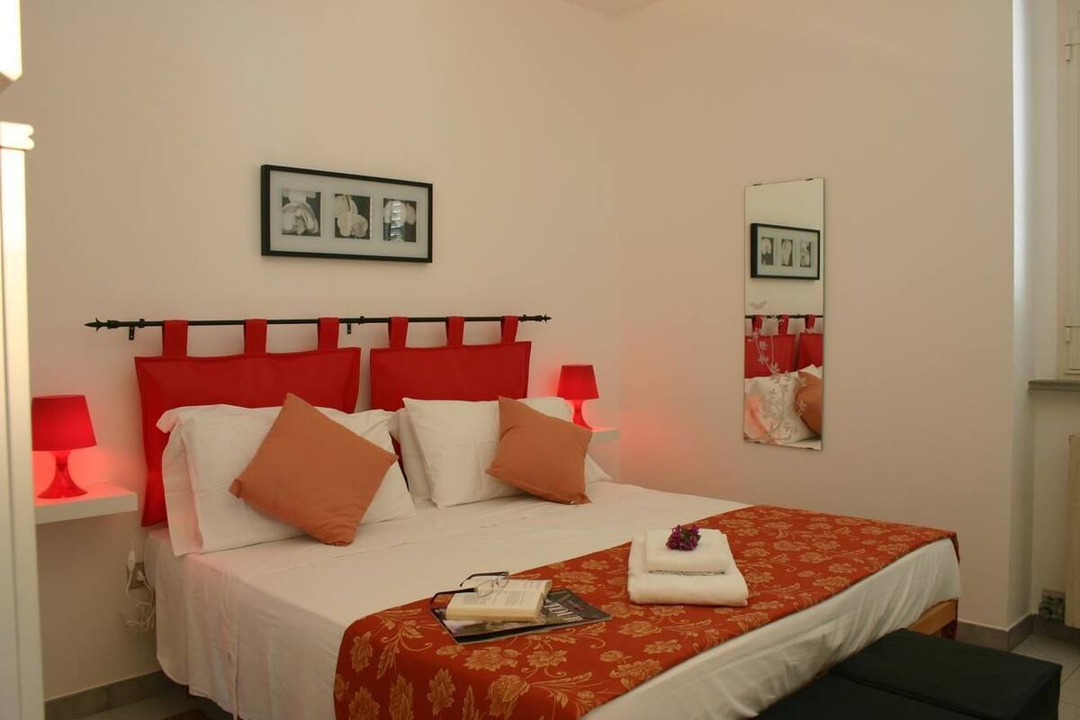 A neatly made bed with a red headboard, white bedding, and orange pillows. There is a bedside table with a red lamp, a framed picture above the bed, and a mirror on the wall.