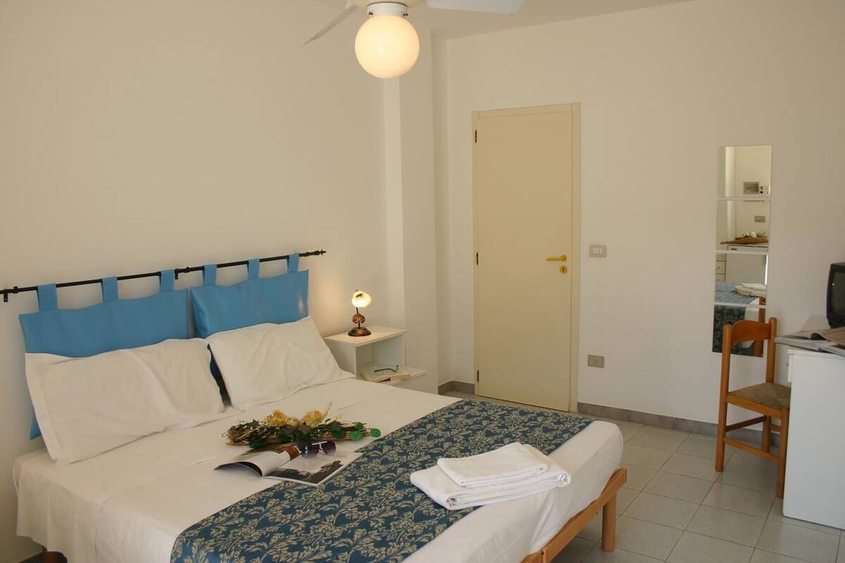 A neatly made bed with a blue and white bedspread, a bedside lamp, and a small table with a lamp. There is a chair and a desk with a computer in the corner.