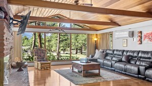 A spacious living room with a leather sofa, wooden coffee table, and a large window offering a view of trees and mountains. - Panorama - Your Private Resort by Estes Park Homes (Estes Park)