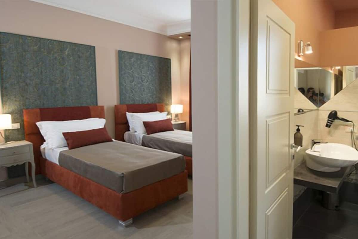 Une chambre d’hôtel avec deux lits, une salle de bain équipée d’un lavabo et d’un miroir, et des œuvres d’art accrochées aux murs.
