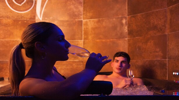 A woman in a bathtub with a man in the background, both holding wine glasses. - Halex Room & Food (Nettuno)