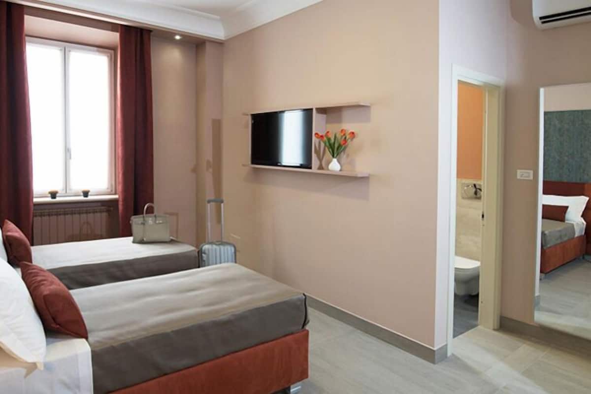Une chambre d’hôtel avec deux lits, une télévision, une petite table ornée d’un vase de fleurs et une salle de bain visible à travers une porte ouverte.