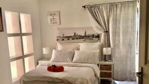 A bedroom with a bed, bedside tables, a wall-mounted light fixture, a framed picture, and a window with curtains. - Beautiful Apartment in Century City (Cape Town)