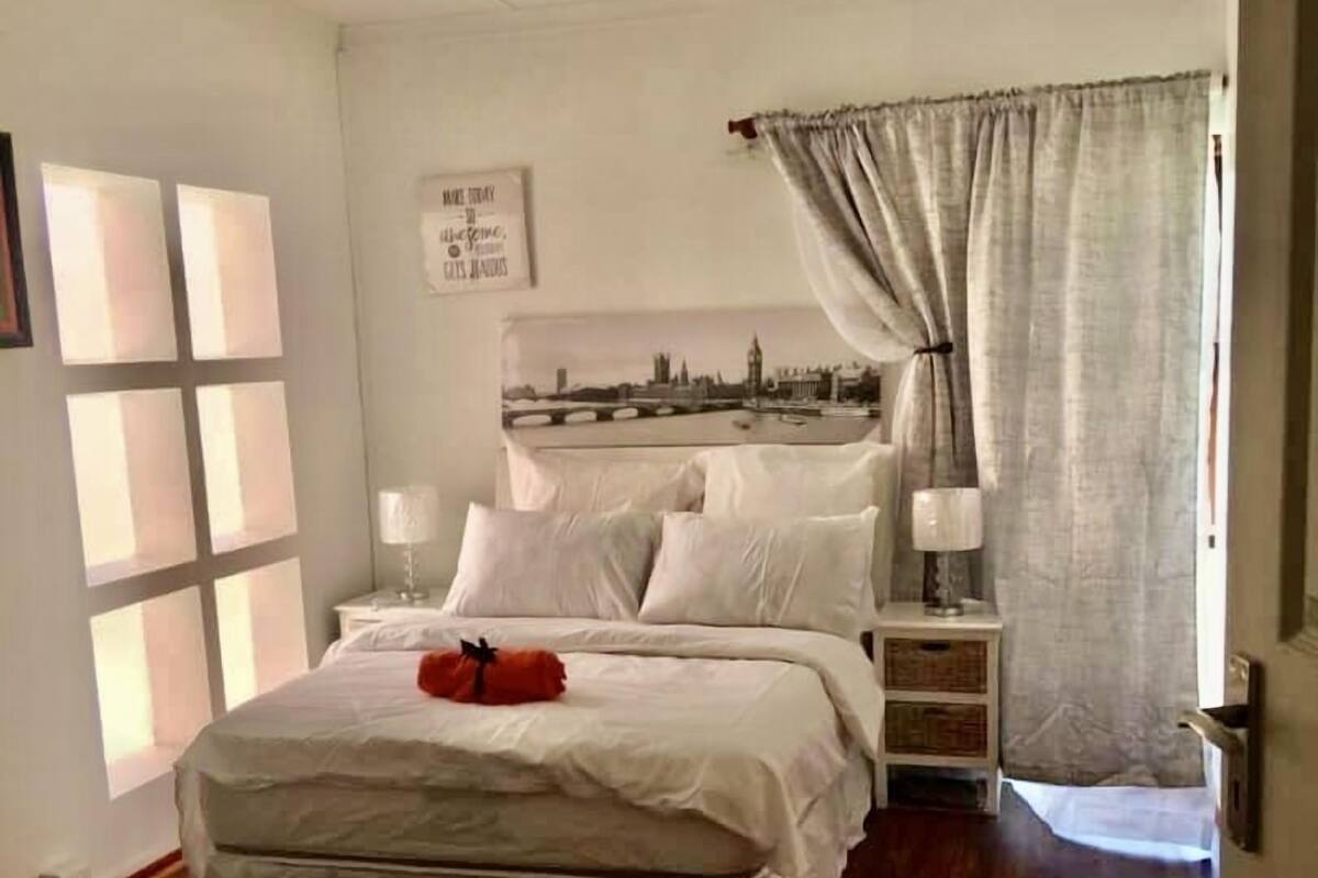 A bedroom with a bed, bedside tables, a wall-mounted light fixture, a framed picture, and a window with curtains.