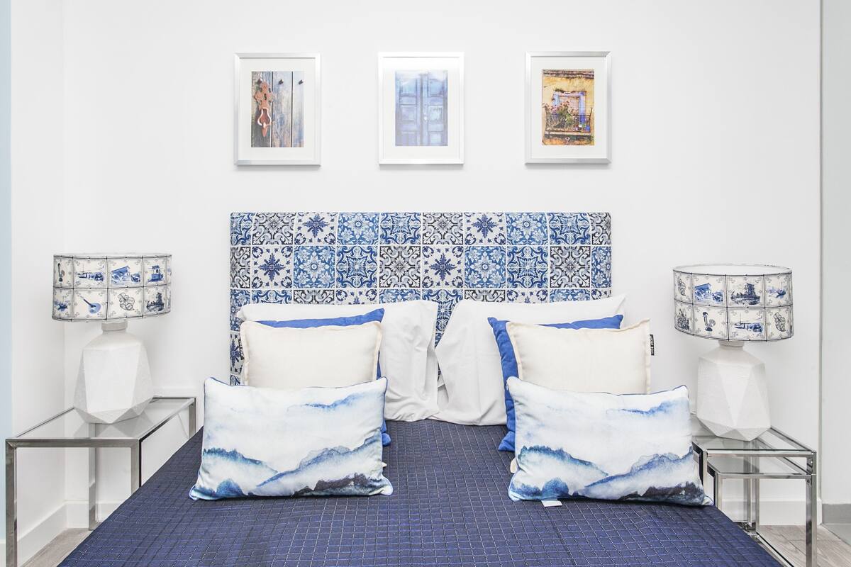 Een slaapkamer met een betegeld hoofdeinde, een bed met blauw-witte beddengoed en twee nachtkastjes met lampen.