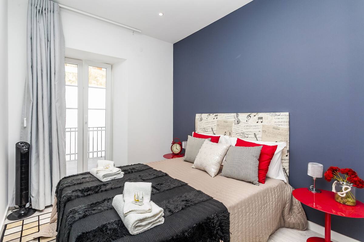 Een slaapkamer met een bed, een rode bijzettafel, een raam met gordijnen en een muur met muzieknoten.