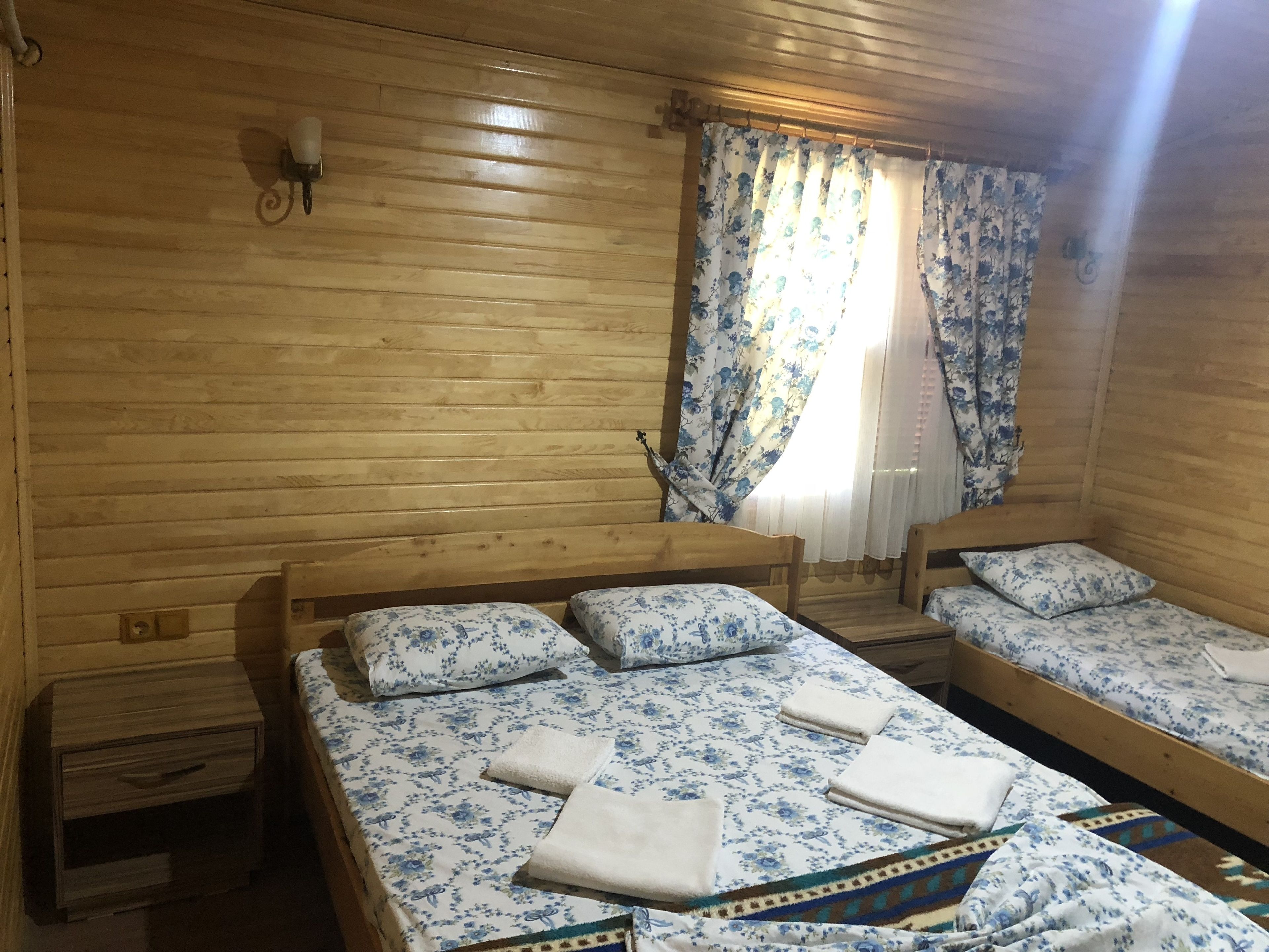 Habitación de cabaña de madera con dos camas, cada una con ropa de cama y almohadas estampadas con motivos florales, una mesita de noche de madera y una ventana con cortinas azules con flores.