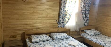 Habitación de cabaña de madera con dos camas, cada una con ropa de cama y almohadas estampadas con motivos florales, una mesita de noche de madera y una ventana con cortinas azules con flores.