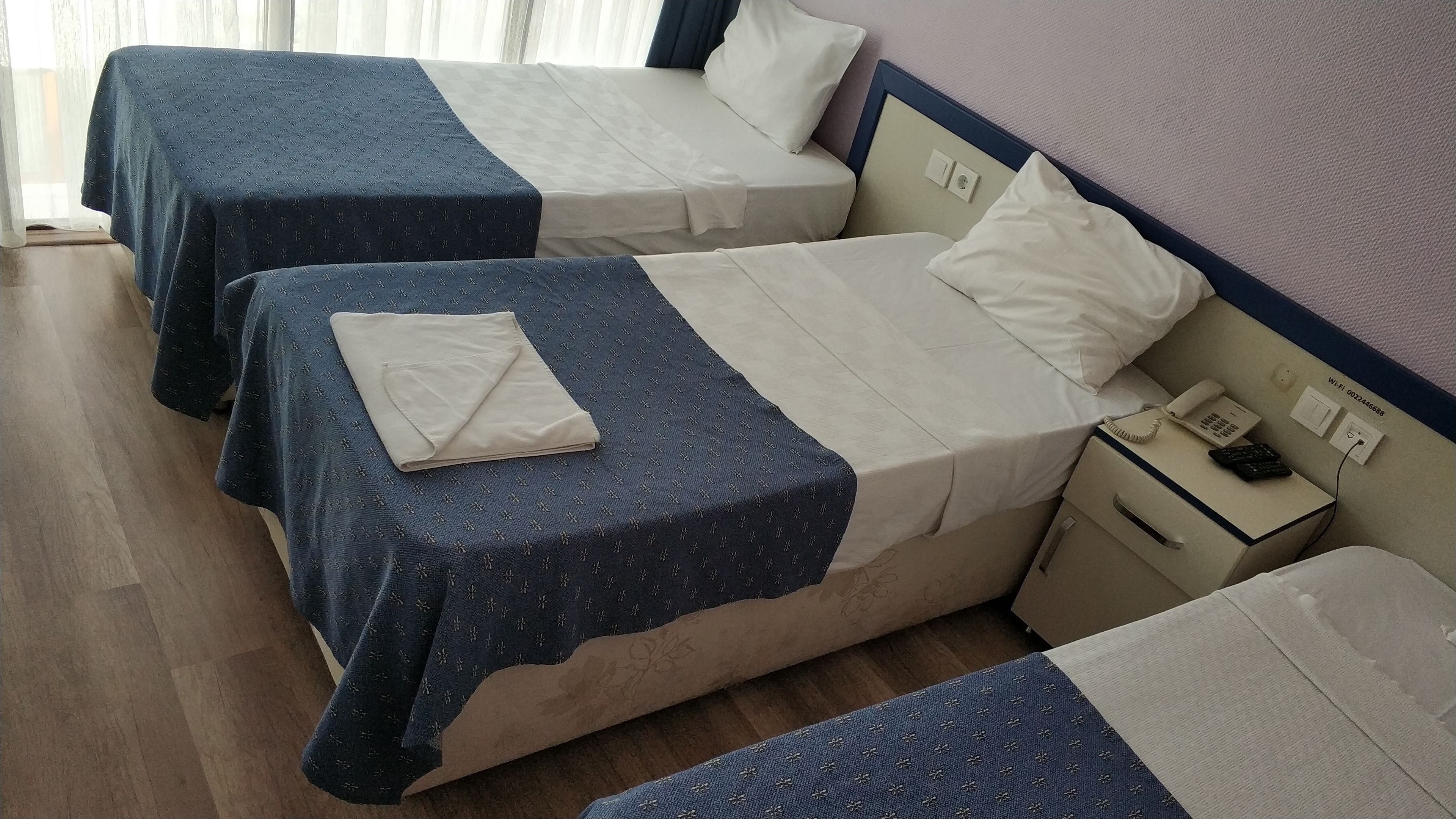 A hotel room with two beds, each with a blue and white bedspread, a bedside table with a phone and a lamp, and a wall-mounted light switch.