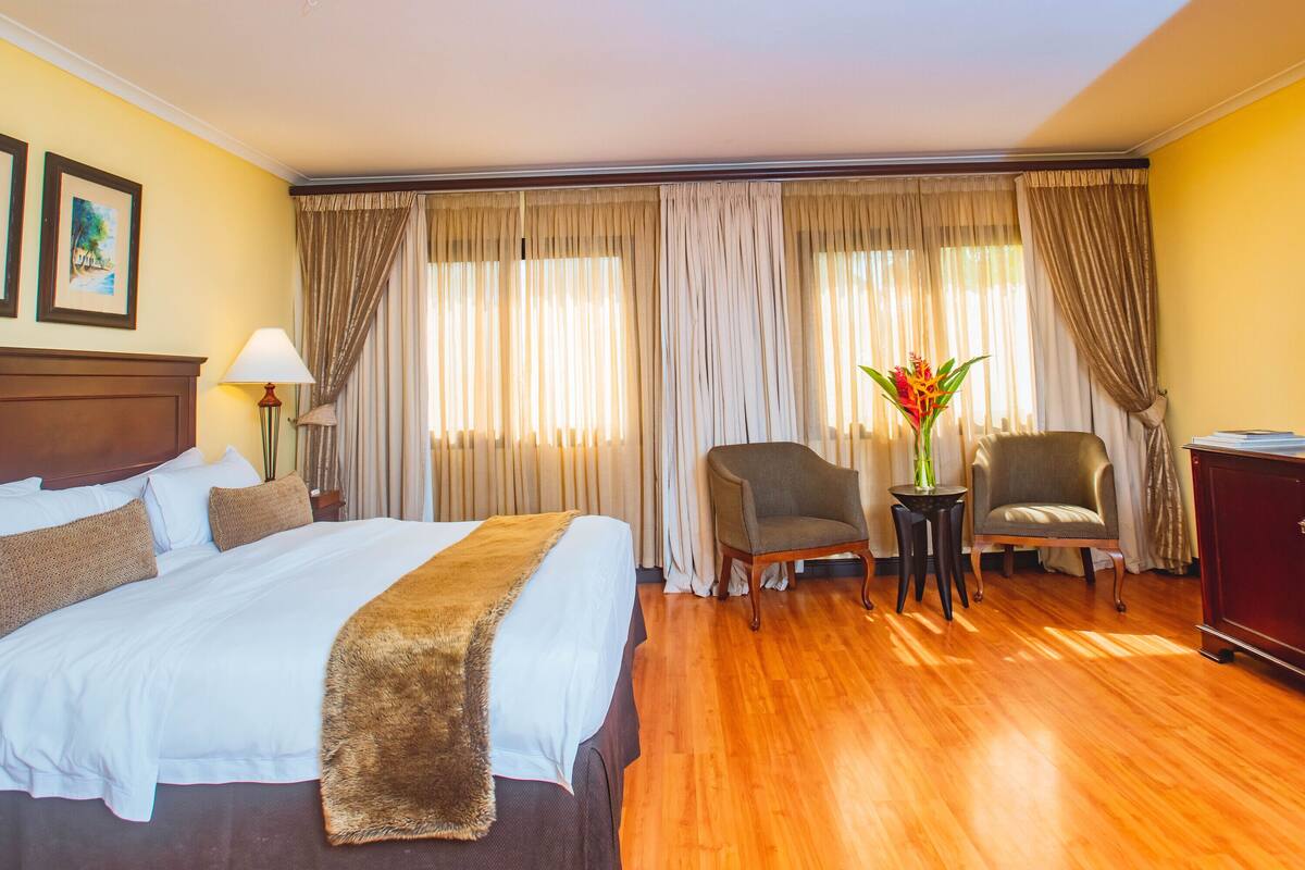 Une chambre d’hôtel avec un grand lit, deux fauteuils, une télévision et un bureau avec une lampe.