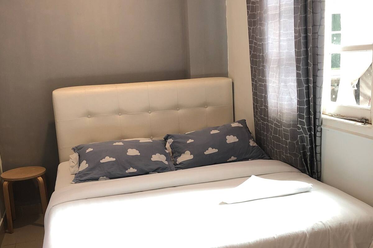 A neatly made bed with a white comforter and two pillows, a small wooden stool, and a window with curtains.