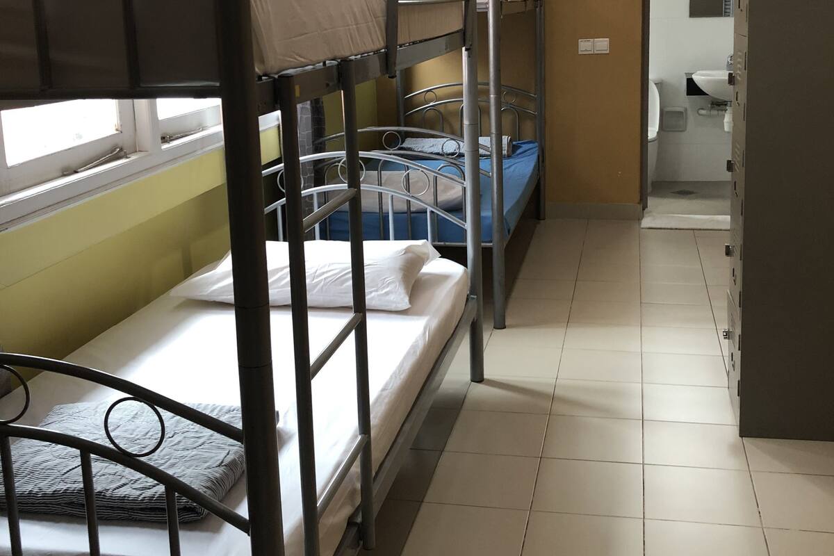 A dormitory room with bunk beds, a window with curtains, a bathroom with a sink and toilet, and a tiled floor.