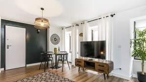 A modern living room with a wooden floor, a dining table with chairs, a large flat-screen TV, a decorative wall piece, and a potted plant. - LxWay Lisboa aos Poiais (Lisbon)