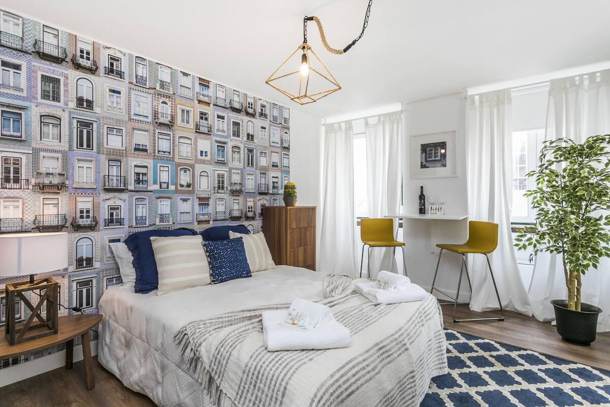 Ein Hotelzimmer mit Wandmuster, einem Bett mit gestreifter Decke, einem Schreibtisch mit zwei Stühlen und einer Topfpflanze.