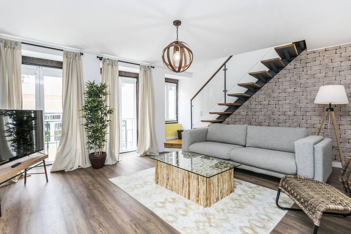Ein modernes Wohnzimmer mit einem grauen Sofa, einem Glastisch, einer Holzbank, einem Blumentopf und einer Treppe an einer Ziegelwand.
