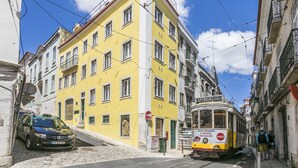 A narrow street with a yellow building, a tram, and a parked car. - LxWay Lisboa aos Poiais (Lisbon)