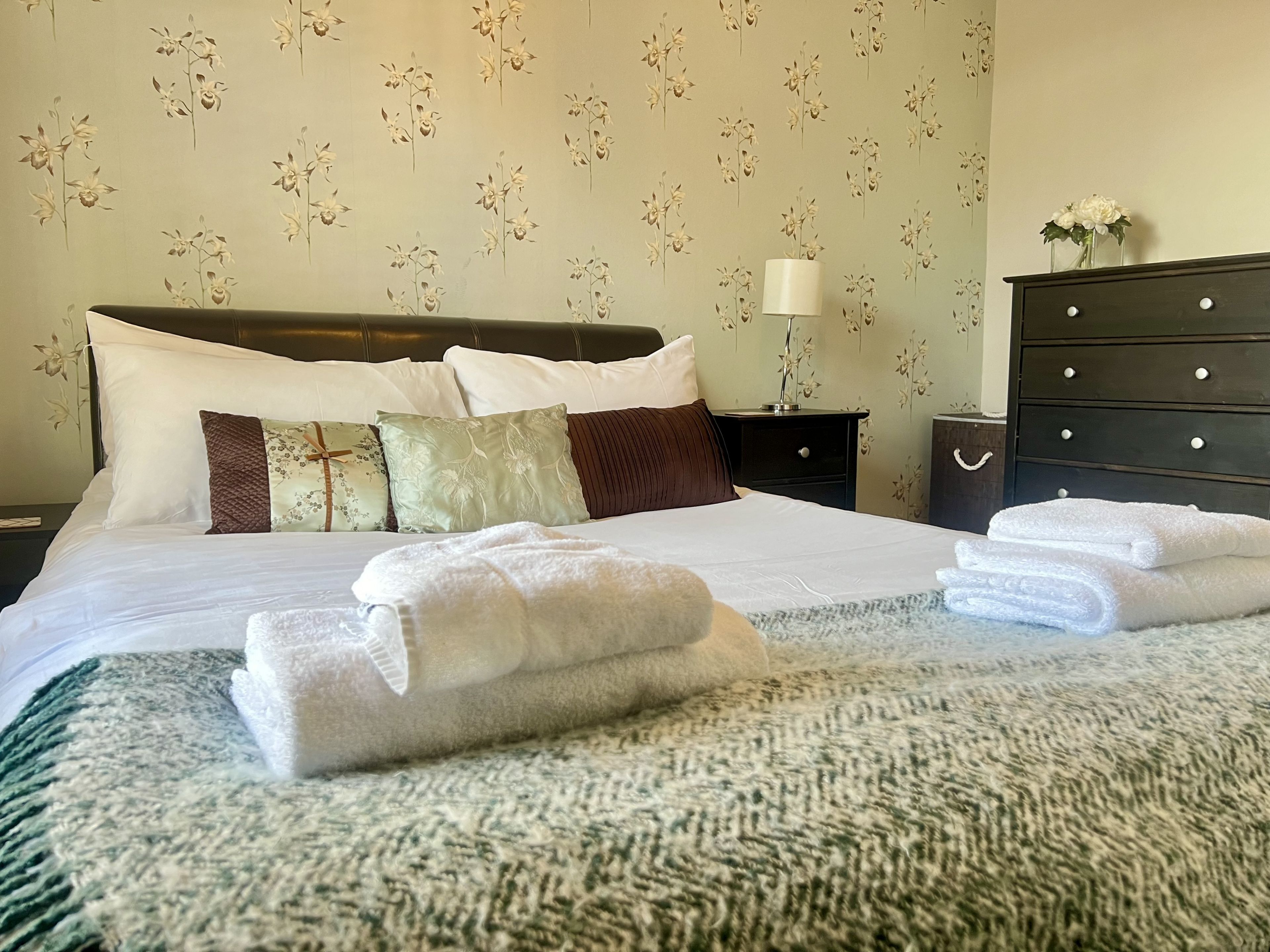 A neatly made bed with white linens and folded towels, a dark wooden dresser, and a nightstand with a lamp.