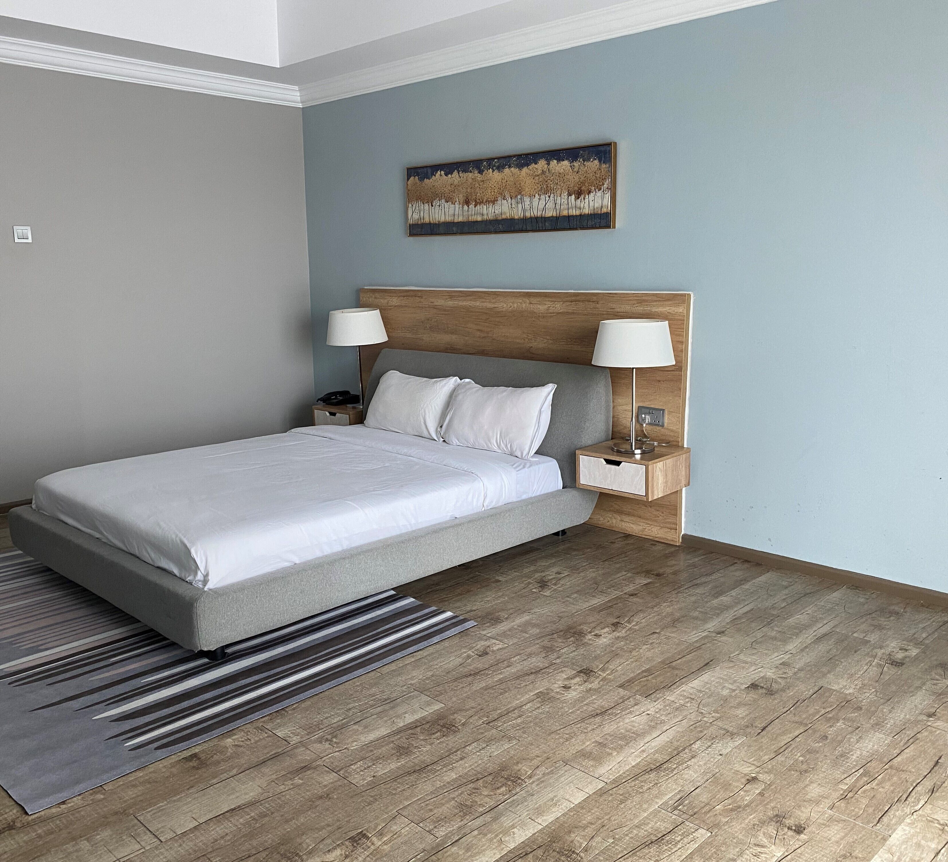 A neatly arranged bedroom with a bed, two bedside tables with lamps, a wooden headboard, a striped rug, and a painting on the wall.