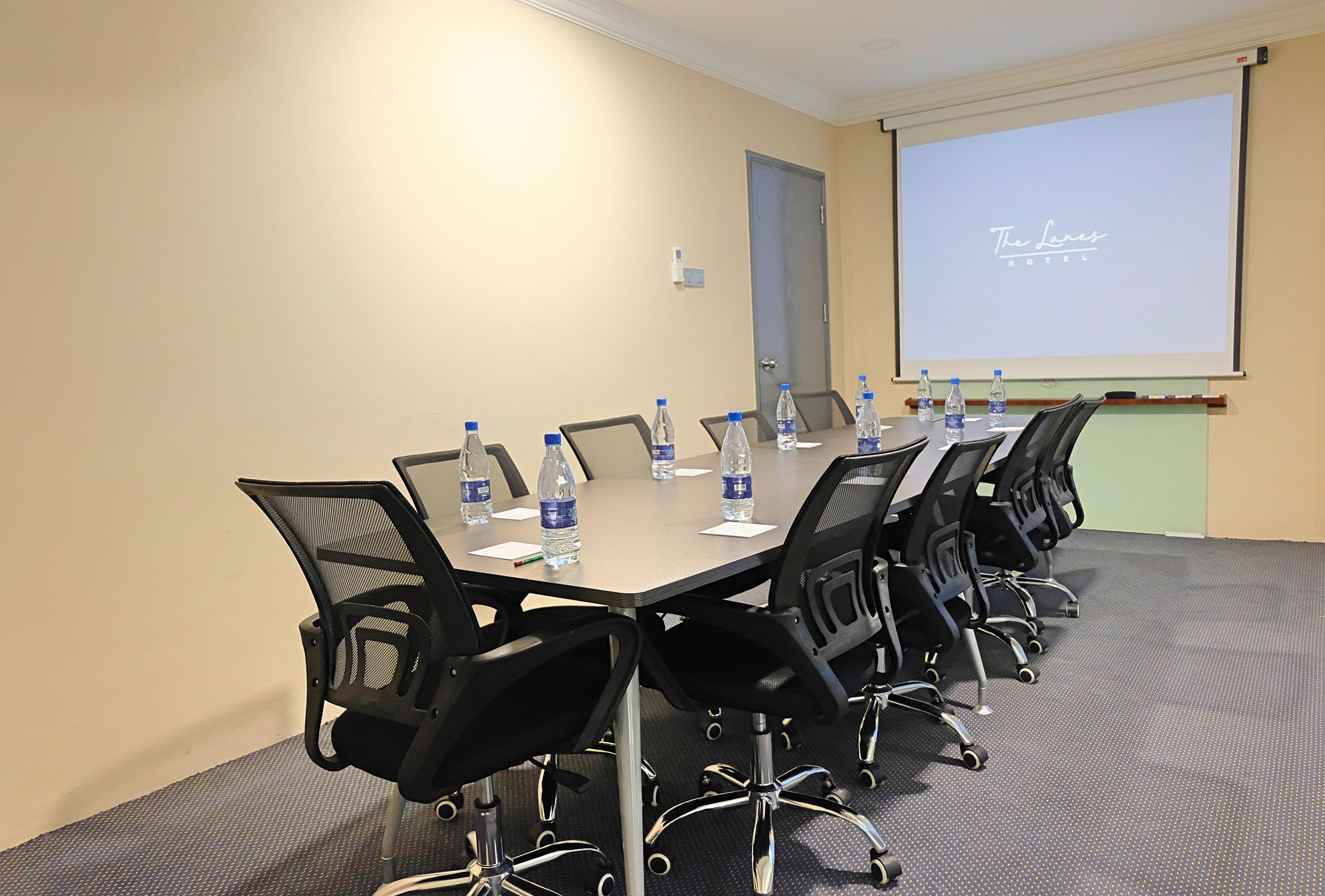 A conference room with a long table, black chairs, water bottles, and a projector screen displaying "The Lane's Hotel" logo.