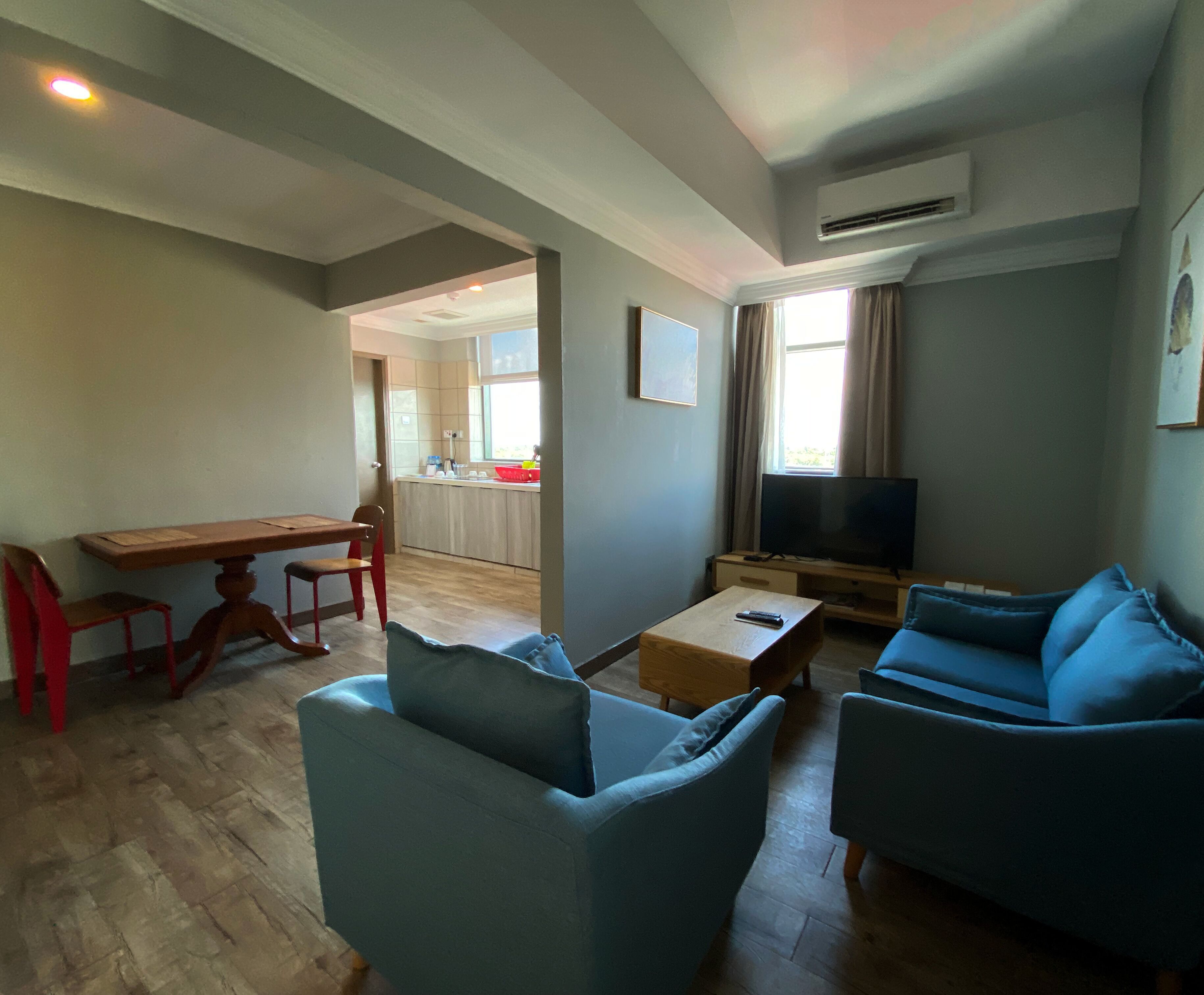 A living room with a TV, two blue sofas, a wooden coffee table, and a dining area with a red chair and a wooden table.