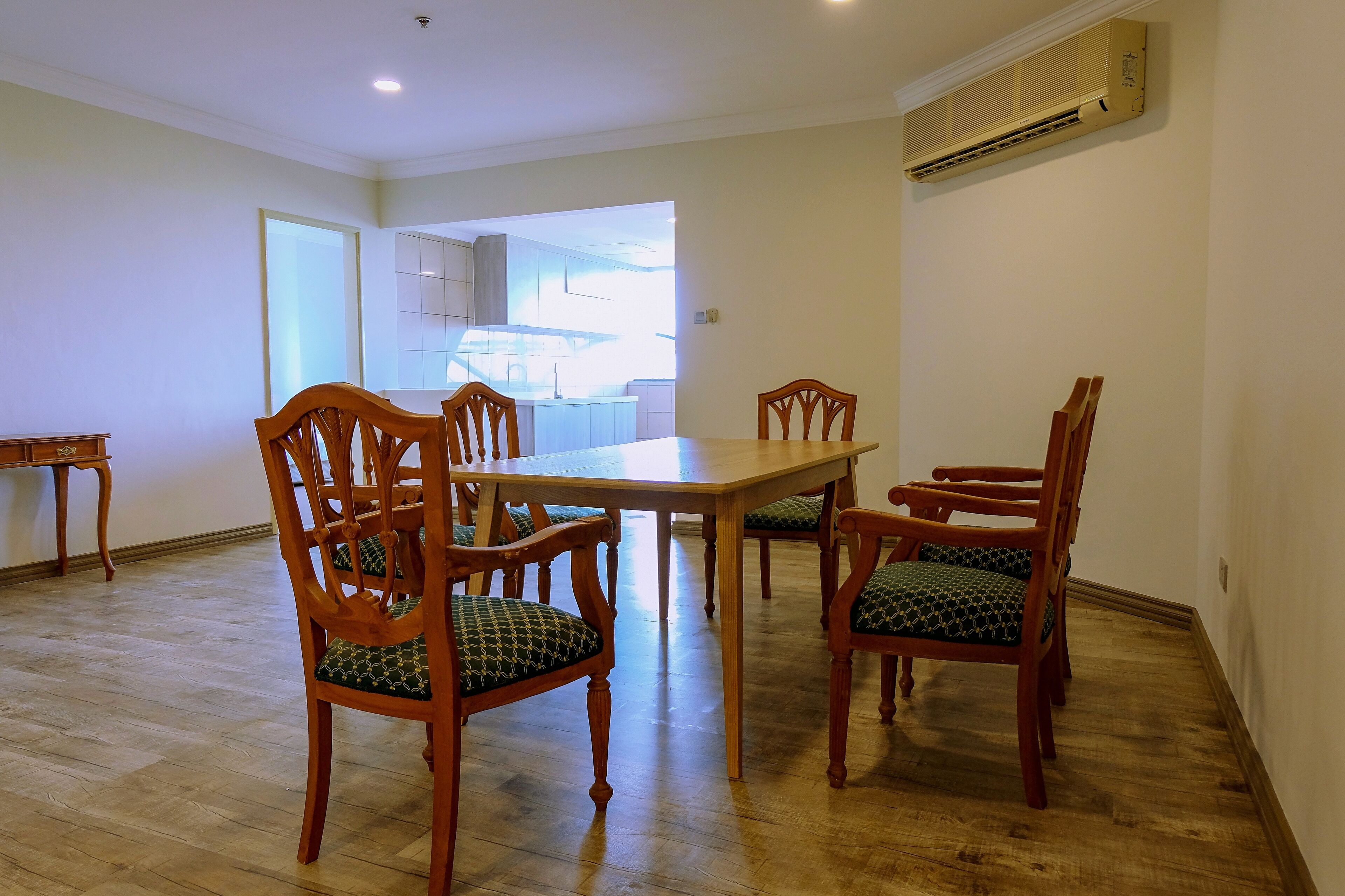 A dining area with a wooden table and chairs, a kitchen in the background, and an air conditioning unit on the wall.