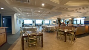 A spacious dining area with round tables and chairs, a central bar, and large windows providing natural light. - The Lanes Hotel (Tutong)