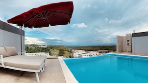 A poolside area with a red umbrella, a white lounge chair, and a view of buildings and hills in the background. - The Lanes Hotel (Tutong)