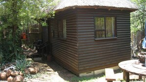 A wooden cabin with a thatched roof, surrounded by trees and a small garden area. - Kruger Inn Backpackers (Nkomazi)