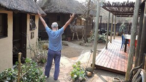 A man standing in a rustic outdoor setting with a wooden structure, a thatched roof, and a wooden deck. - Kruger Inn Backpackers (Nkomazi)
