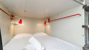 Un letto rifatto con lenzuola bianche e un asciugamano arrotolato, due lampade rosse fissate al muro e un tubo rosso che corre lungo il soffitto.