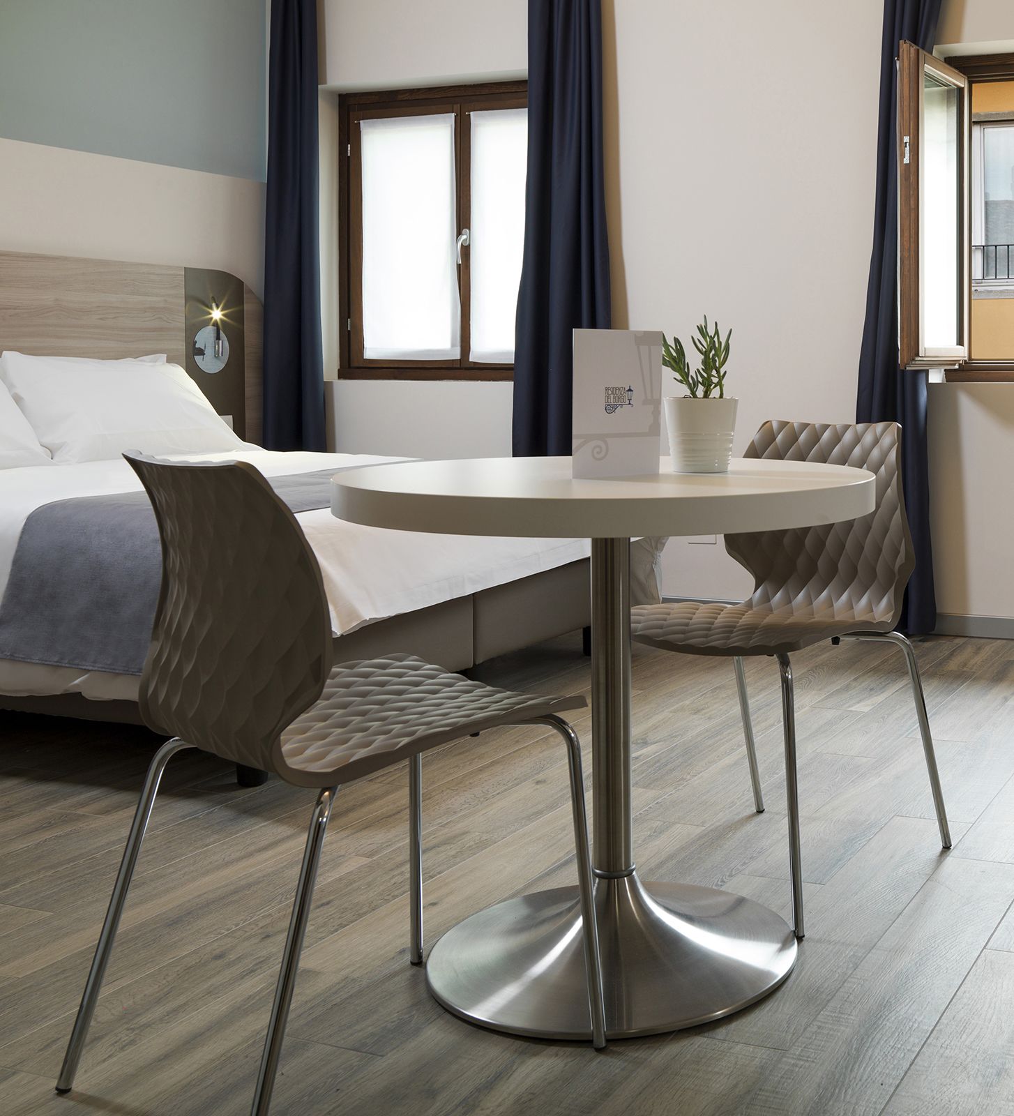 A modern hotel room with a bed, a round table with two chairs, a potted plant, and a window with curtains.