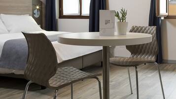 A modern hotel room with a bed, a round table with two chairs, a potted plant, and a window with curtains.
