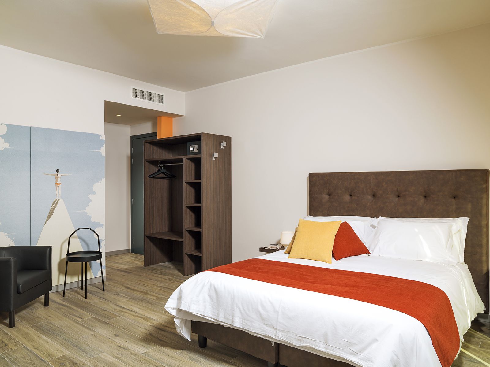 A hotel room with a bed, a wardrobe, a chair, and a wall with a painting.