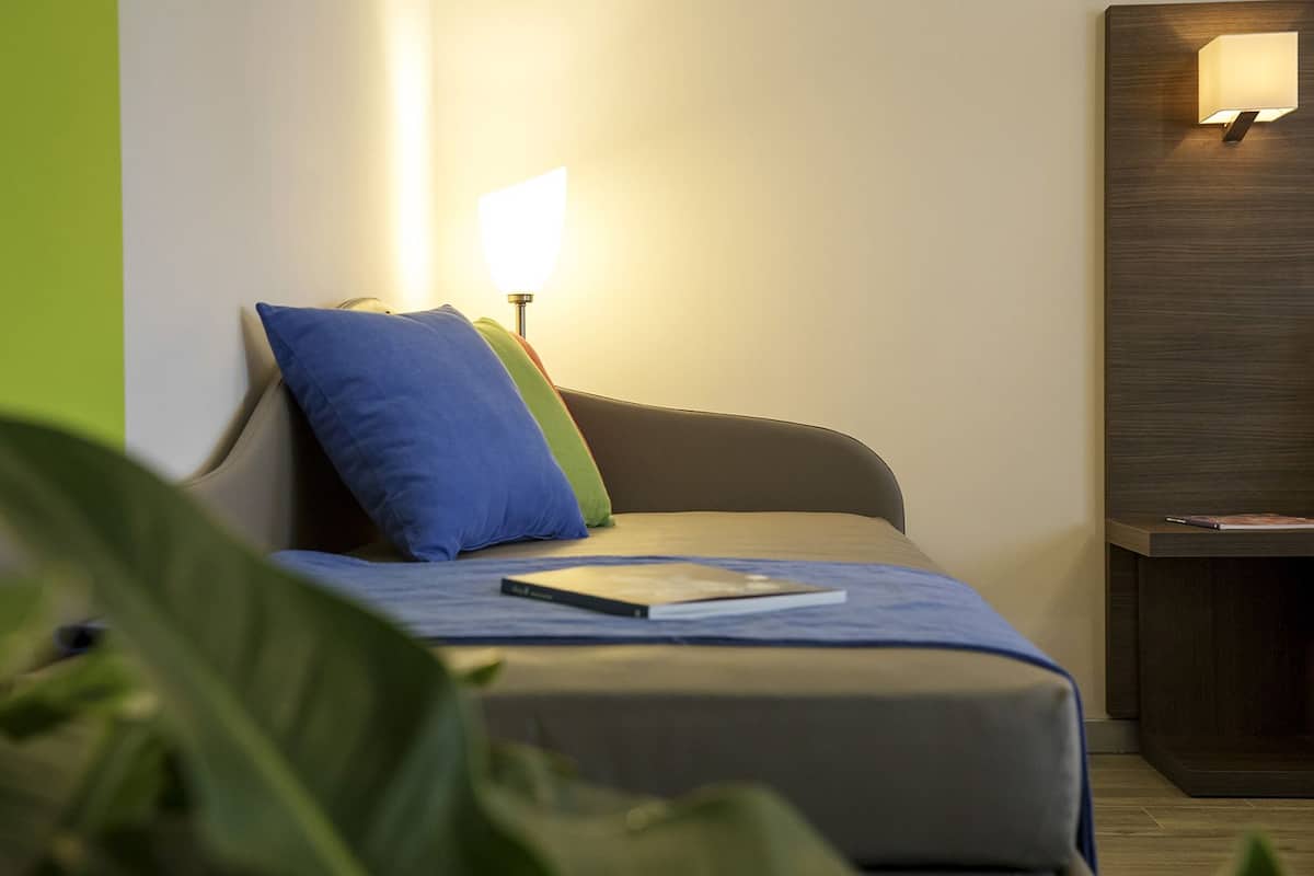 Un letto rifatto con un cuscino blu e uno verde, una lampada da comodino e un tavolino con un libro.