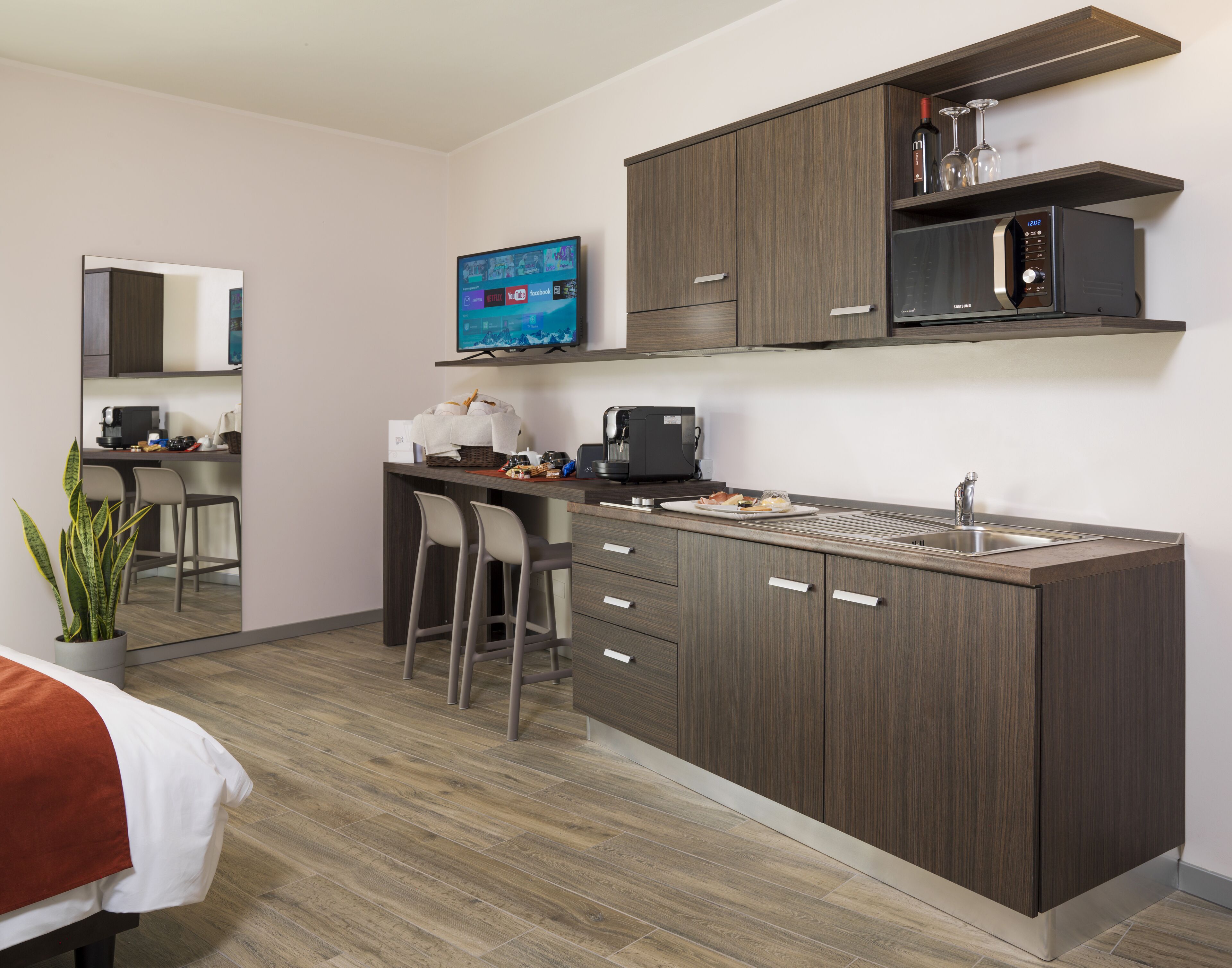 A modern kitchen with a built-in microwave, a coffee machine, and a sink. There are bar stools and a small dining area with a TV mounted on the wall.