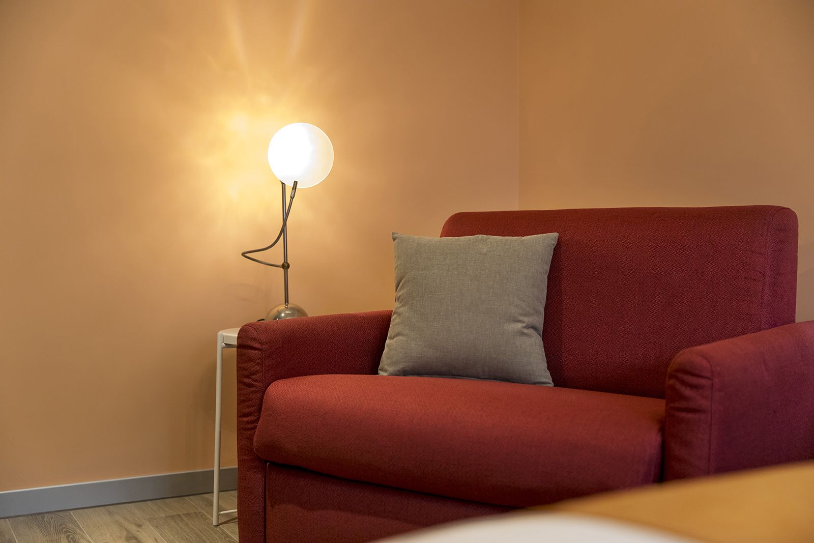 A red armchair with a grey pillow against a plain wall, next to a floor lamp with a round light.