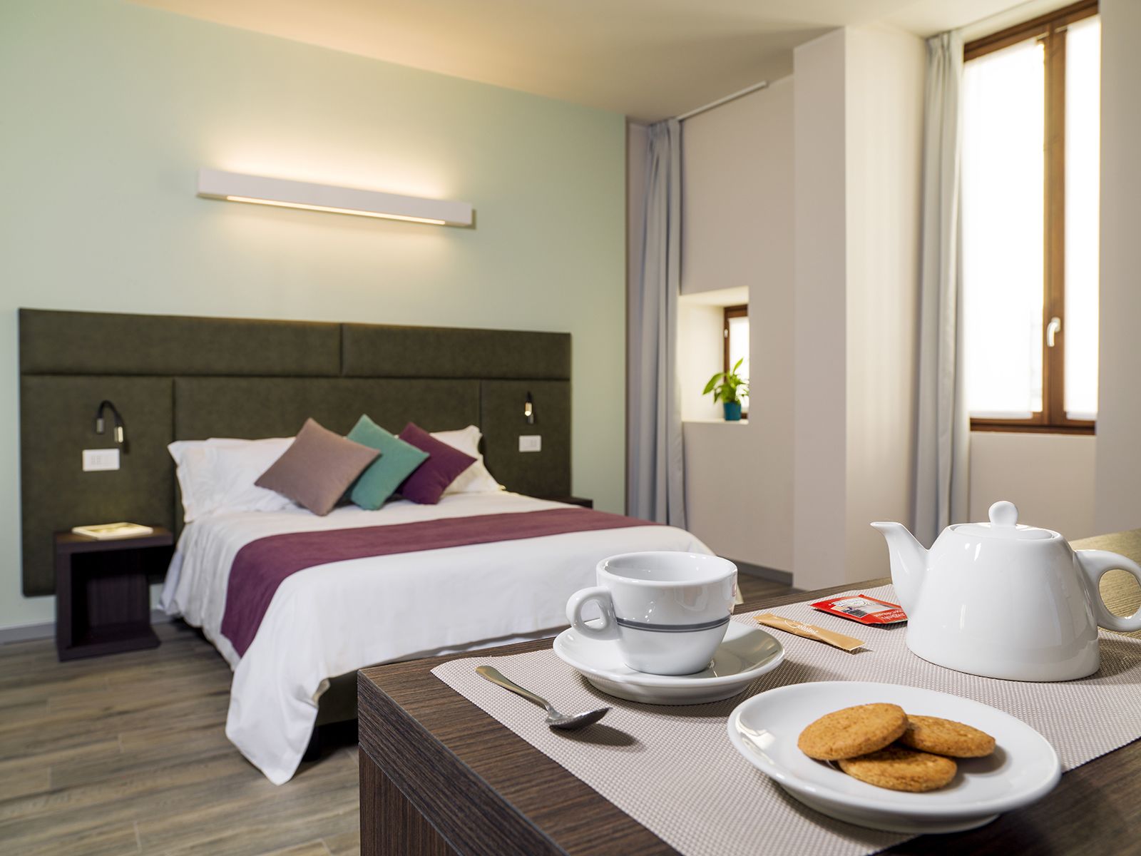A hotel room with a neatly made bed, a bedside table with a cup, a teapot, and cookies, and a window with curtains.