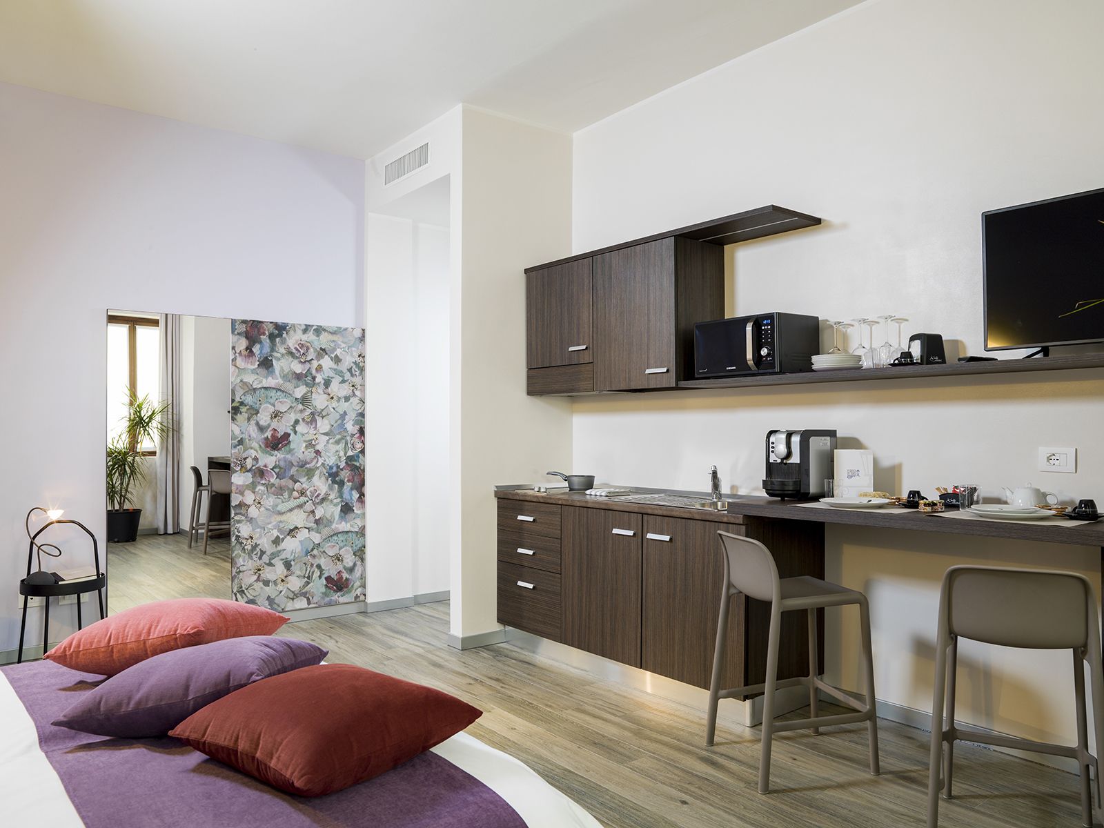 A modern kitchen with dark wooden cabinets, a microwave, and a coffee maker. There is a dining area with a table and chairs, and a bed with pillows.