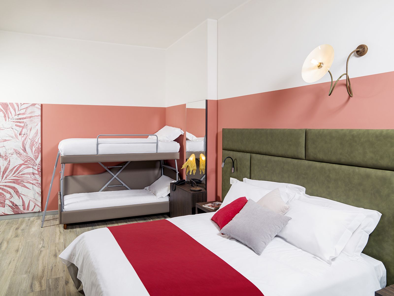 Habitación de hotel con literas, una cama individual con cubrecama rojo y blanco, una mesita de noche con lámpara, y un aplique de pared.