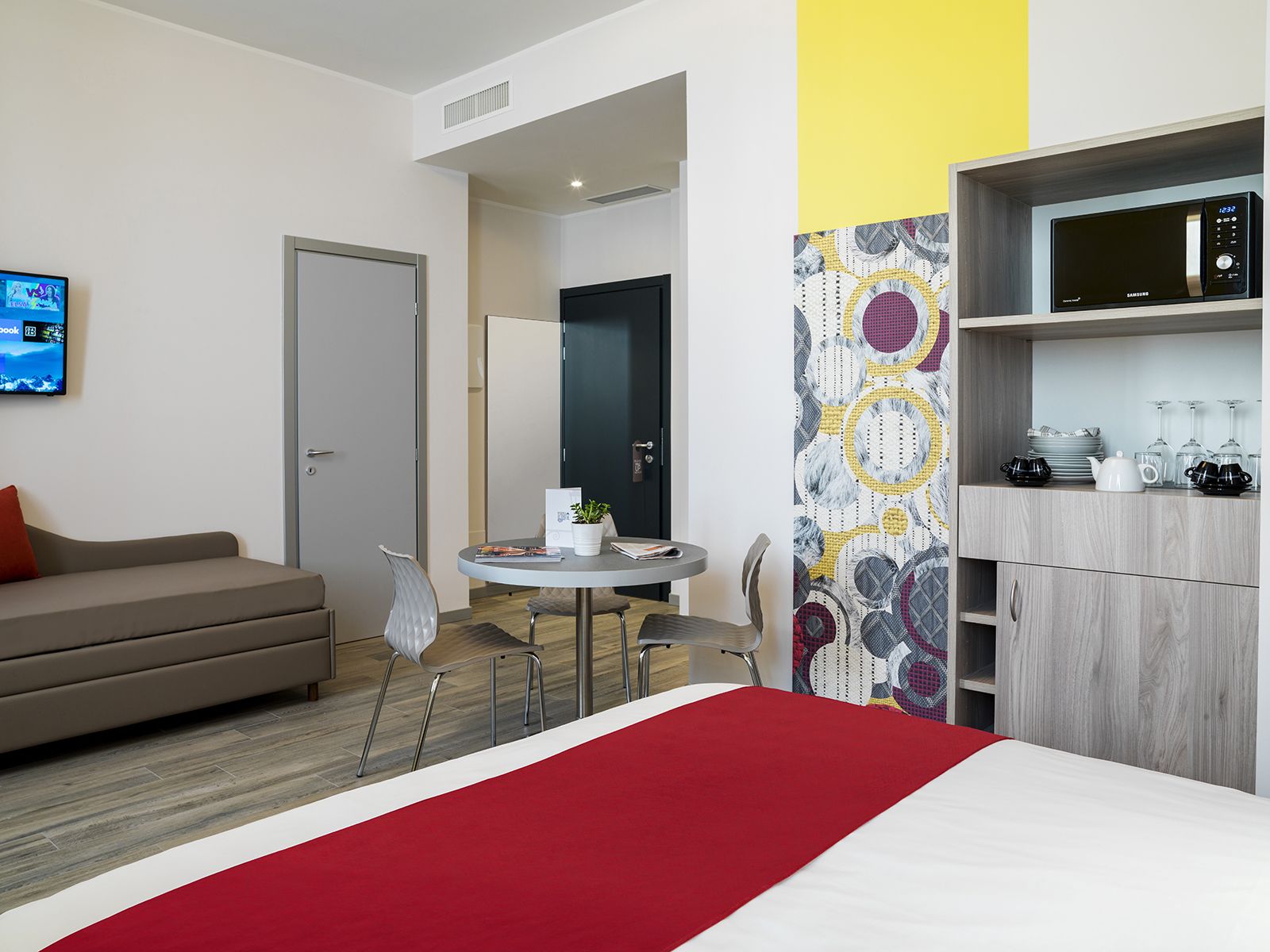 A modern hotel room with a bed, a sofa, a dining table with chairs, a TV, a microwave, and a patterned wall.